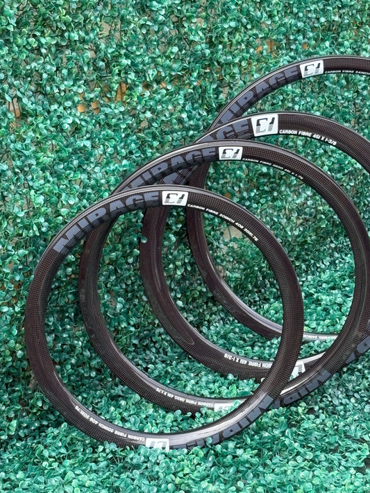 Four black carbon BMX bicycle tires with white Mirage sidewall branding arranged in a circle on a green textured artificial turf background.